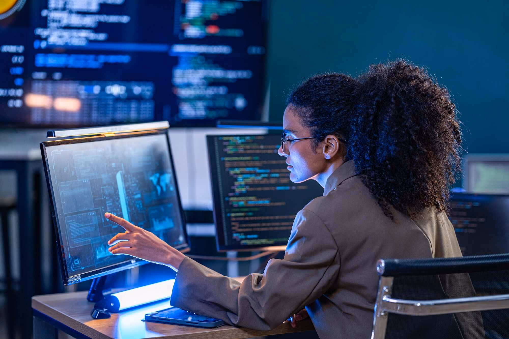 A woman with curly hair and glasses in a brown jacket is seated at a desk, pointing at a computer screen displaying digital data and graphics, with code visible on nearby monitors in a dimly lit room.