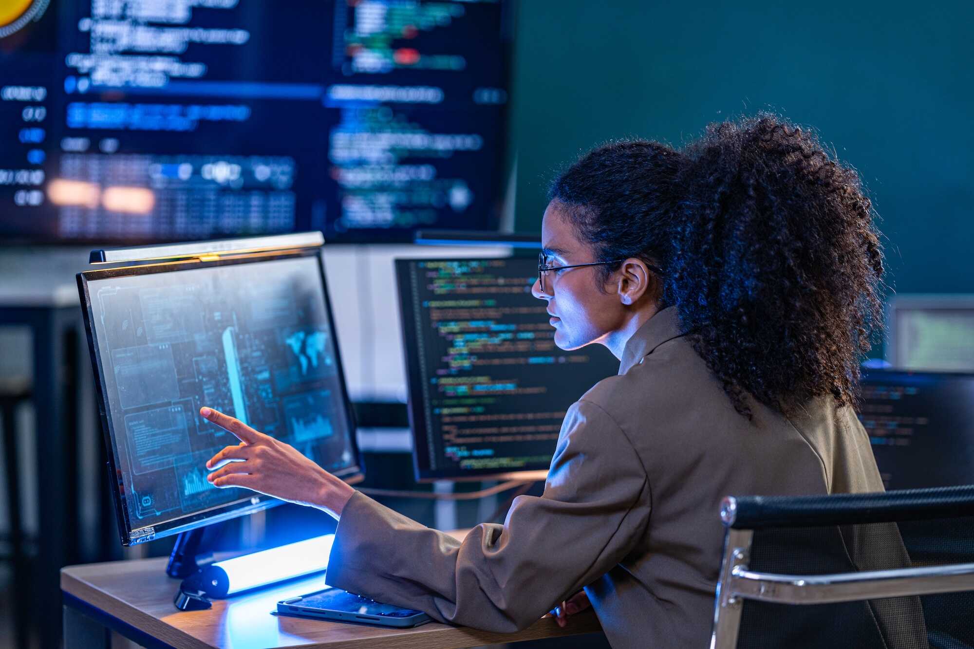 A woman with curly hair and glasses in a brown jacket is seated at a desk, pointing at a computer screen displaying digital data and graphics, with code visible on nearby monitors in a dimly lit room.