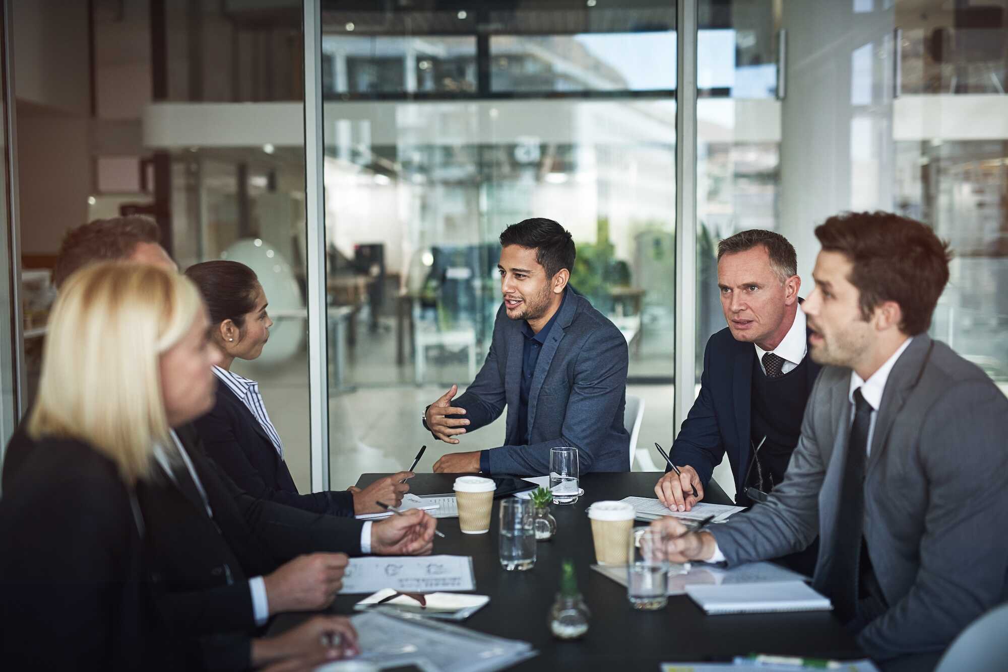 Six business professionals having a meeting around a table in a modern office with glass walls, engaged in discussion with papers, notebooks, and coffee cups on the table.