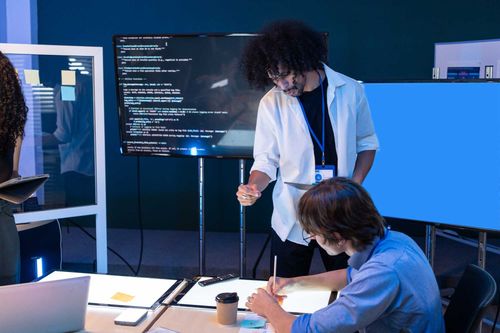 Two people collaborating in a tech workspace with multiple screens, one showing code, the other blue, a man pointing while another writes at a lighted desk with a coffee cup and notes.