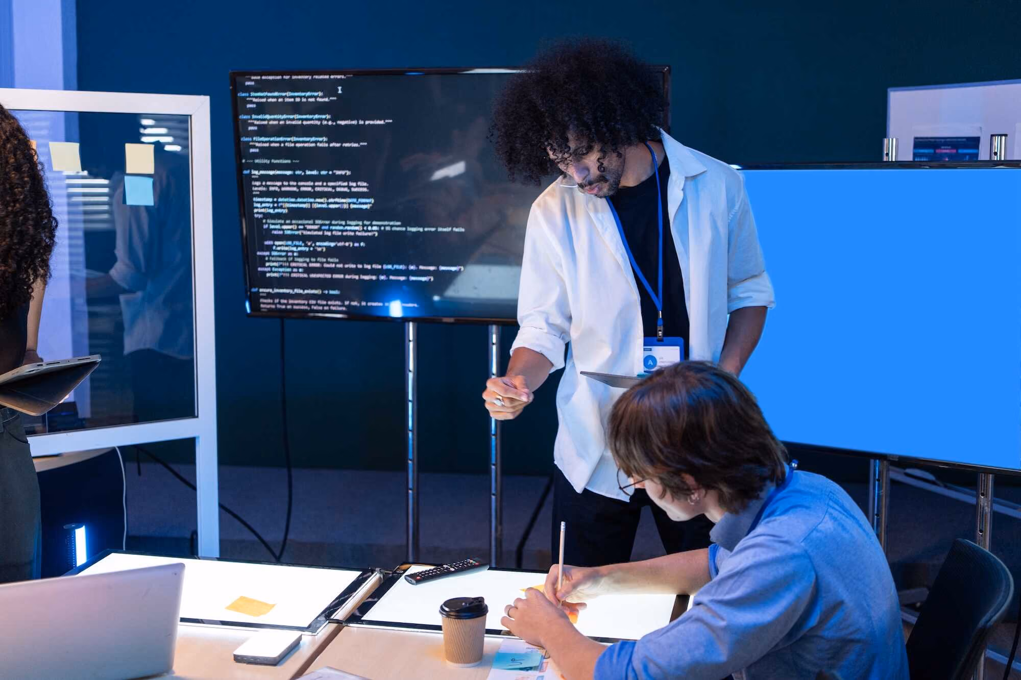 Two people collaborating in a tech workspace with multiple screens, one showing code, the other blue, a man pointing while another writes at a lighted desk with a coffee cup and notes.