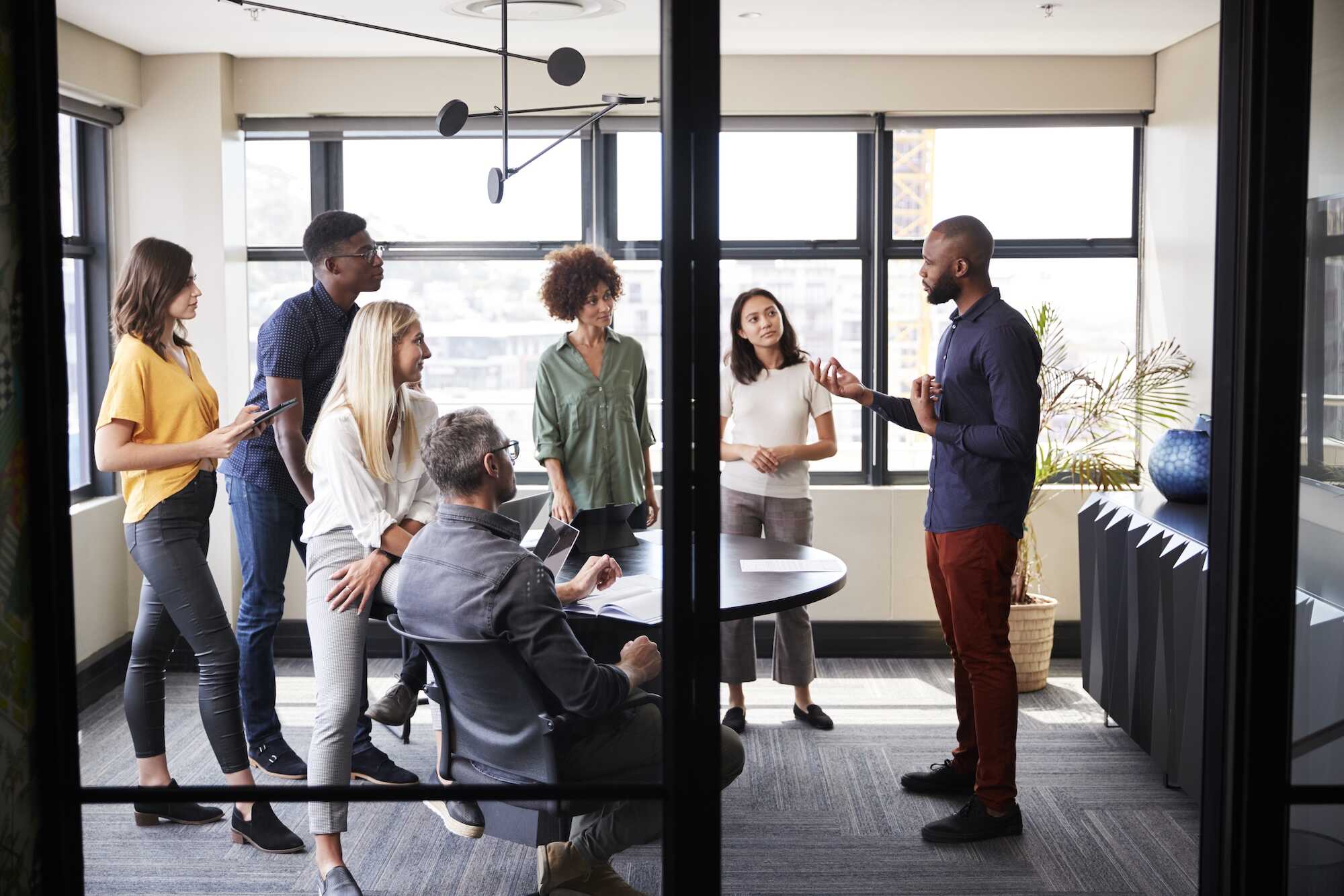 A diverse group of six professionals in a modern office meeting room, with five people standing and one seated around a table, attentively listening to a man speaking and gesturing.