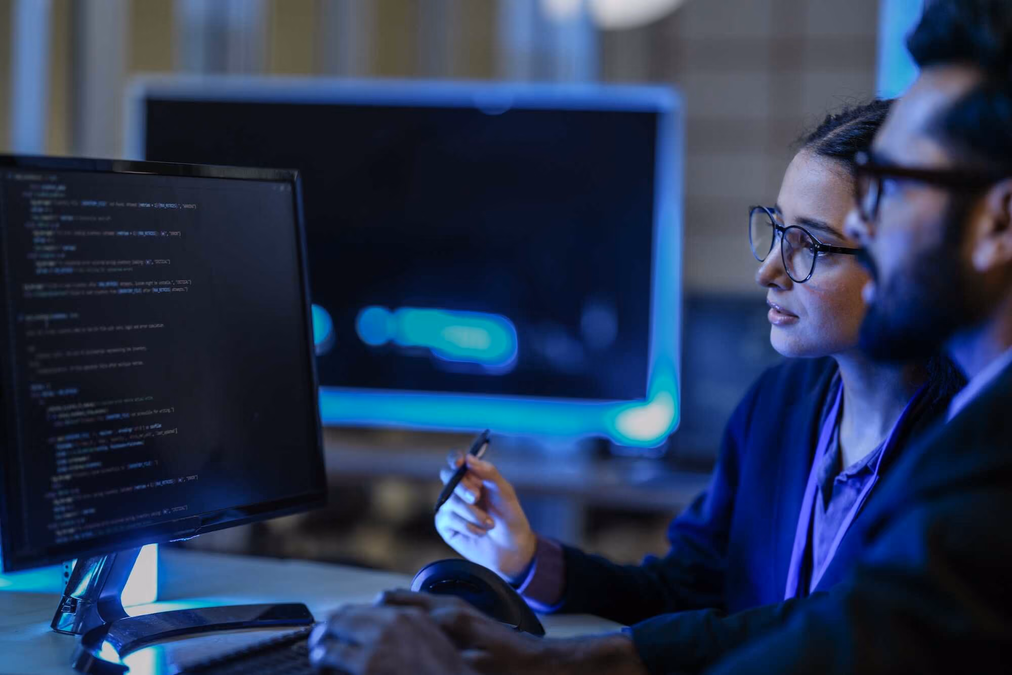 Two people with glasses looking at a computer screen displaying programming code in a dimly lit office.
