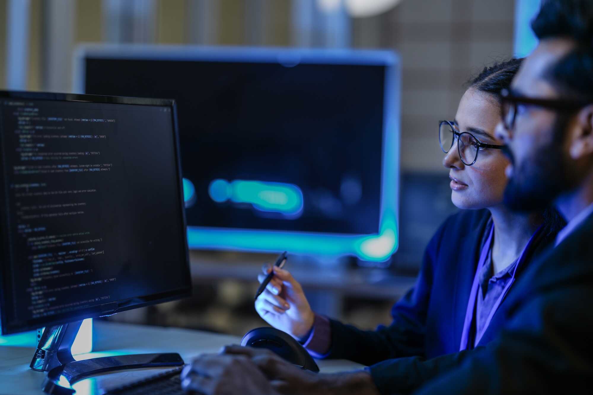 Two people with glasses looking at a computer screen displaying programming code in a dimly lit office.