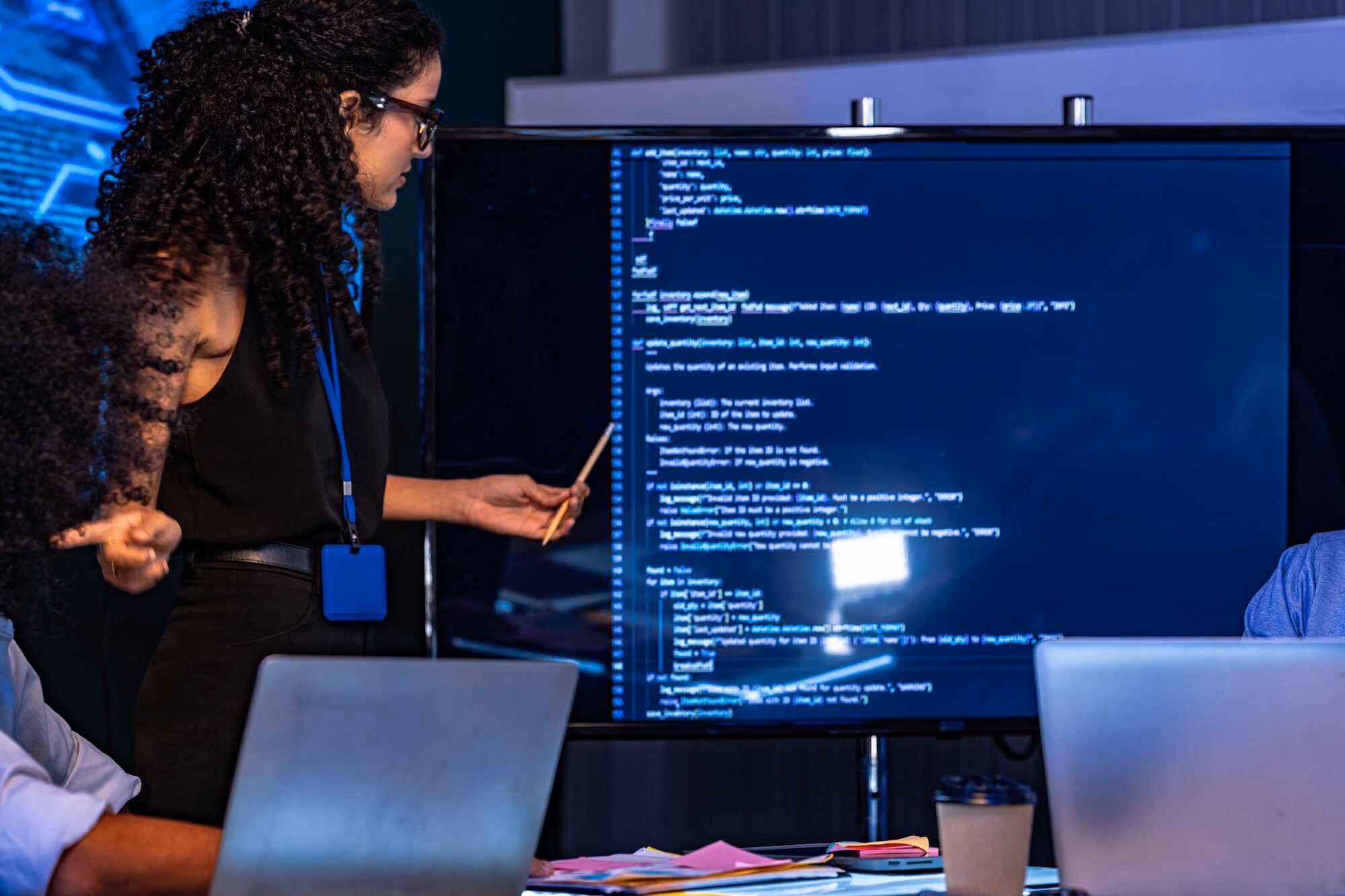 A woman with curly hair and glasses pointing at a large screen displaying computer code, while two people with laptops are partially visible seated around a table with papers and a coffee cup, in a tech workspace setting.