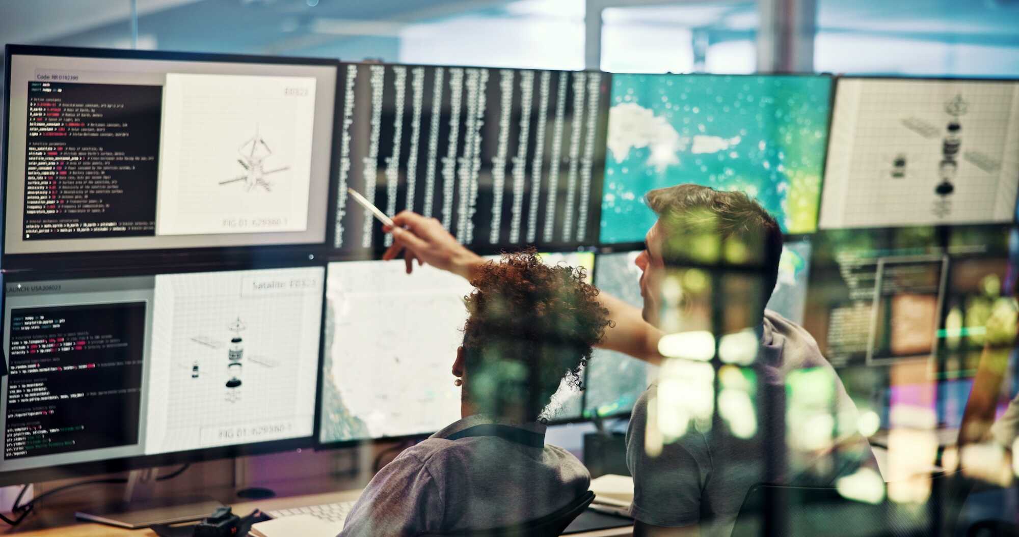 Two people in a control room with multiple large monitors displaying code, satellite diagrams, and data; one person points at a screen while discussing with the other.
