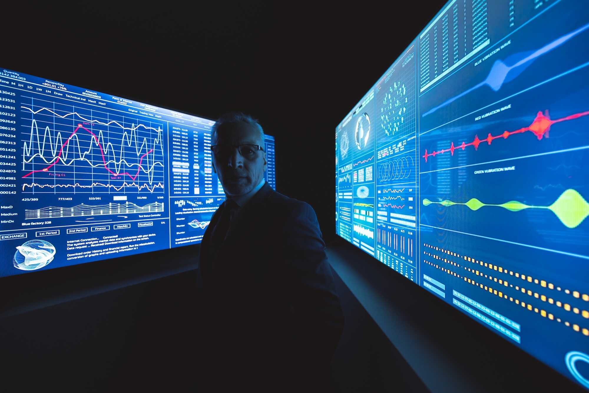 Man in suit standing in a dark room between two large digital screens displaying complex data, charts, and graphs in blue light.