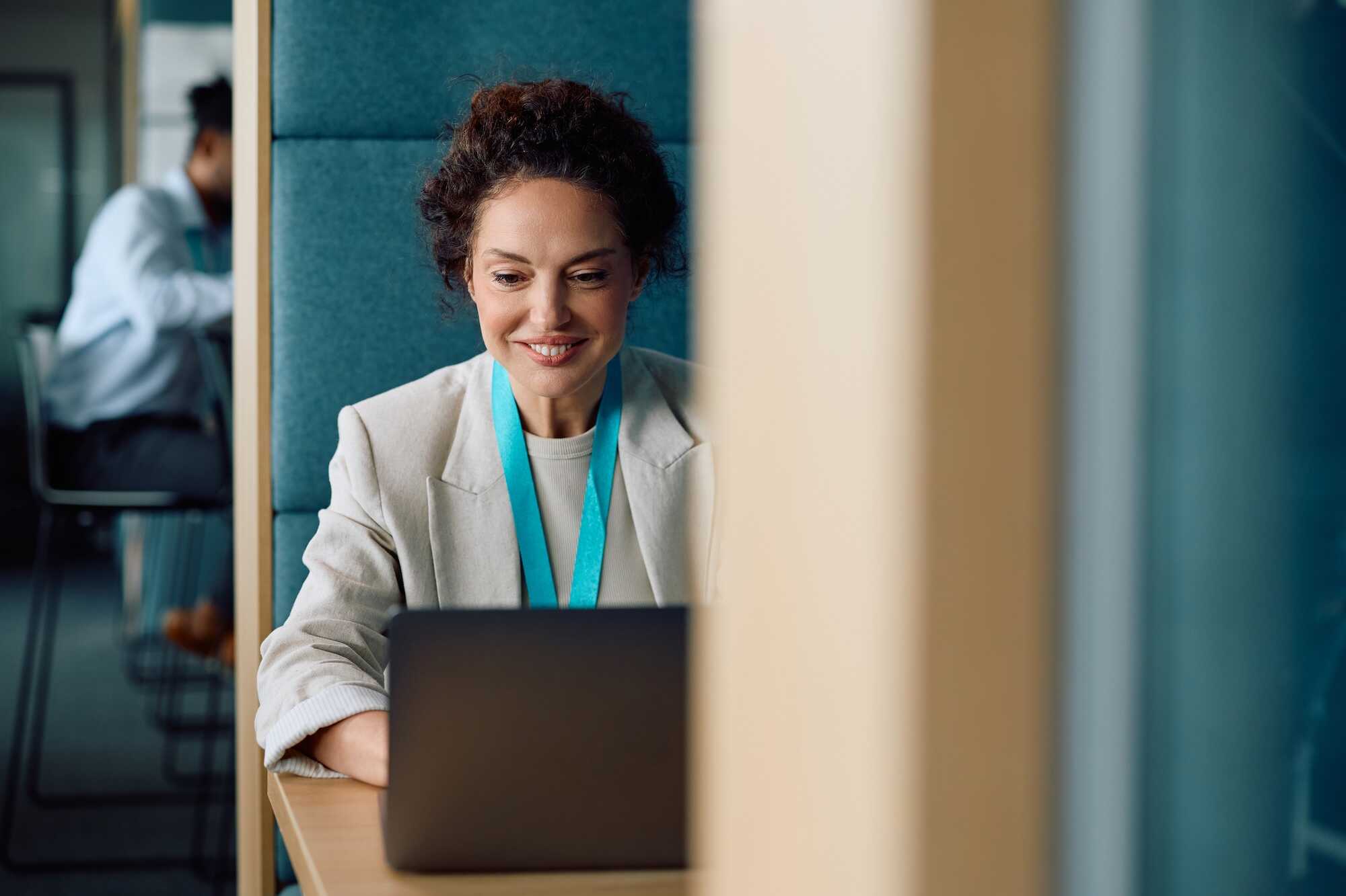 Smiling woman with curly hair and a beige blazer working on a laptop at a wooden desk in an office booth, wearing a blue lanyard.