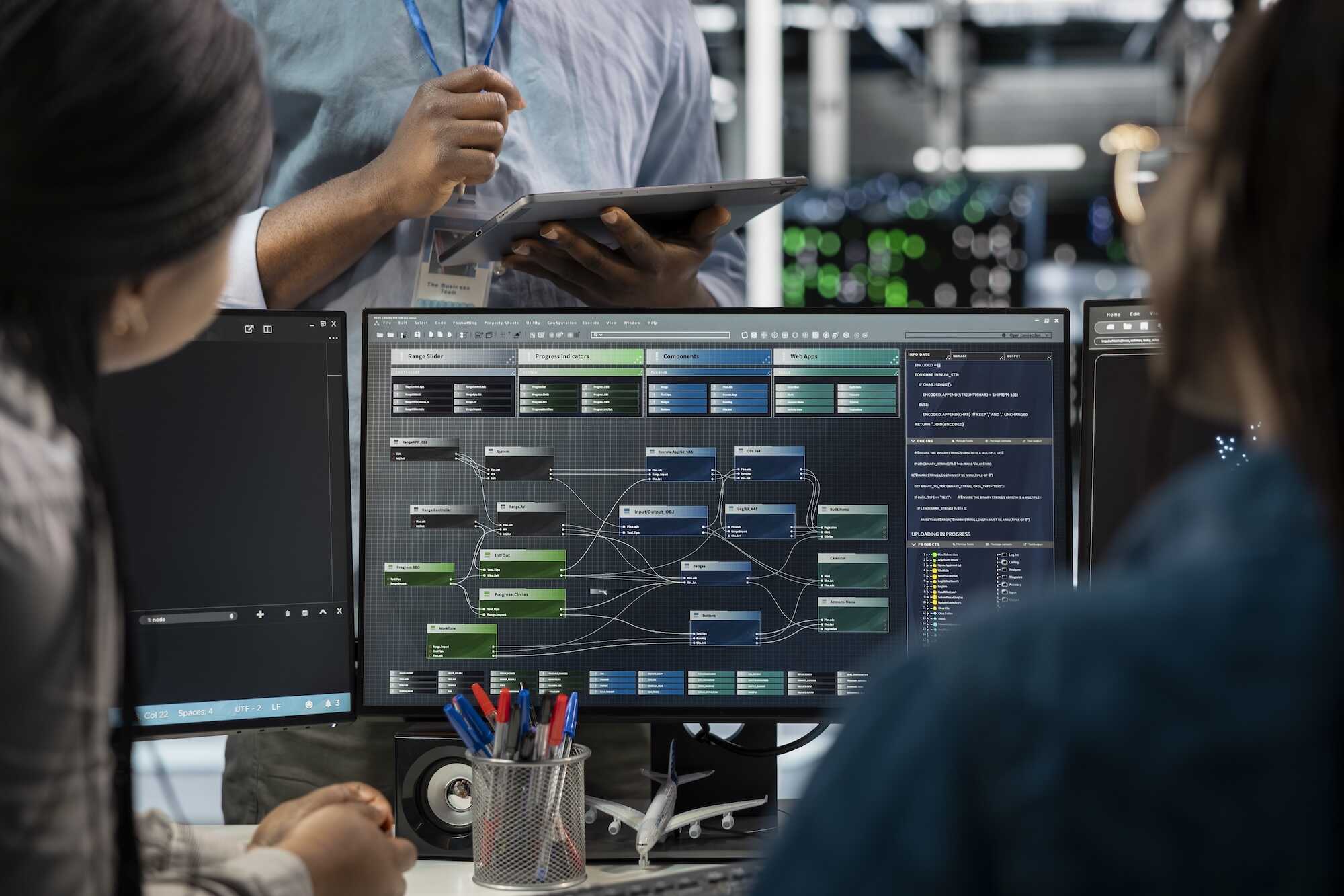 Two people looking at a computer screen displaying a software interface with interconnected modules labeled Range Slider, Progress Indicators, Components, and Web Apps, while a third person holds a tablet in the background in an office setting.