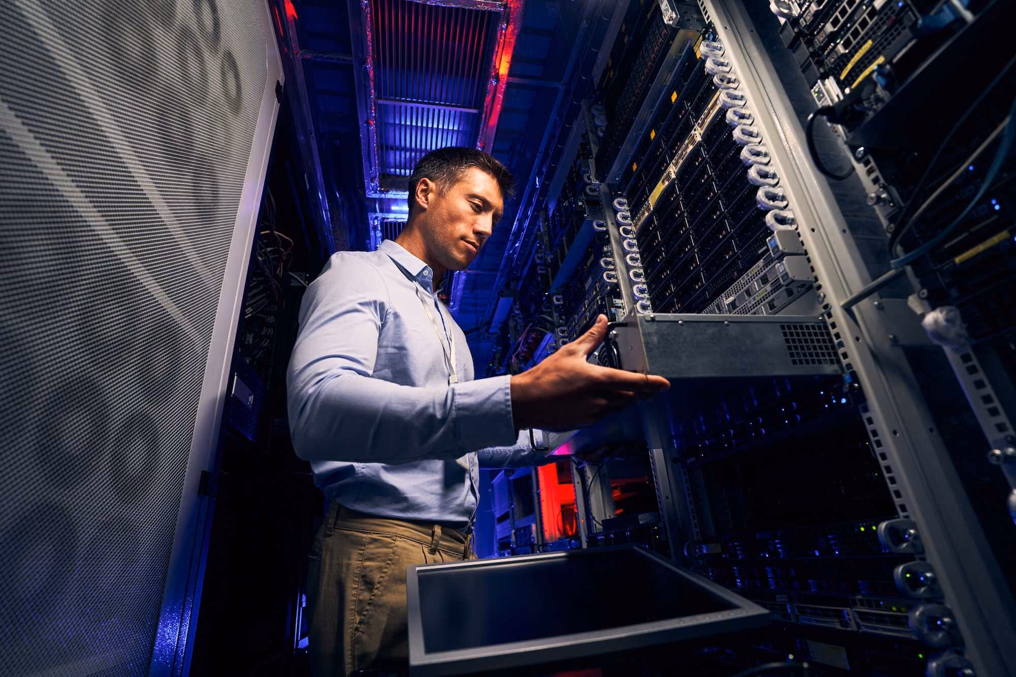 A man in a light blue shirt lifts a server unit out from a rack inside a dimly lit data center with blue and red lighting.