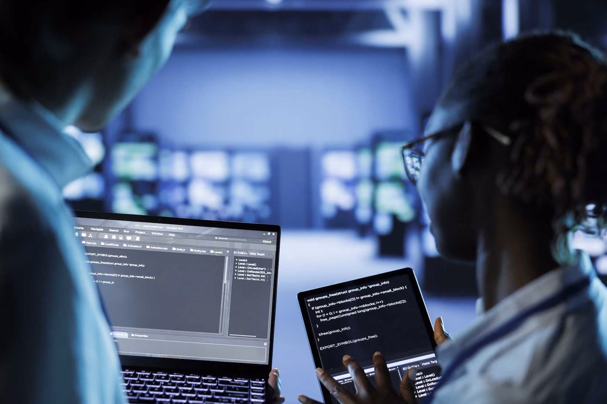 Two programmers in a dimly lit room working together, one holding a laptop and the other a tablet, both displaying computer code on their screens.