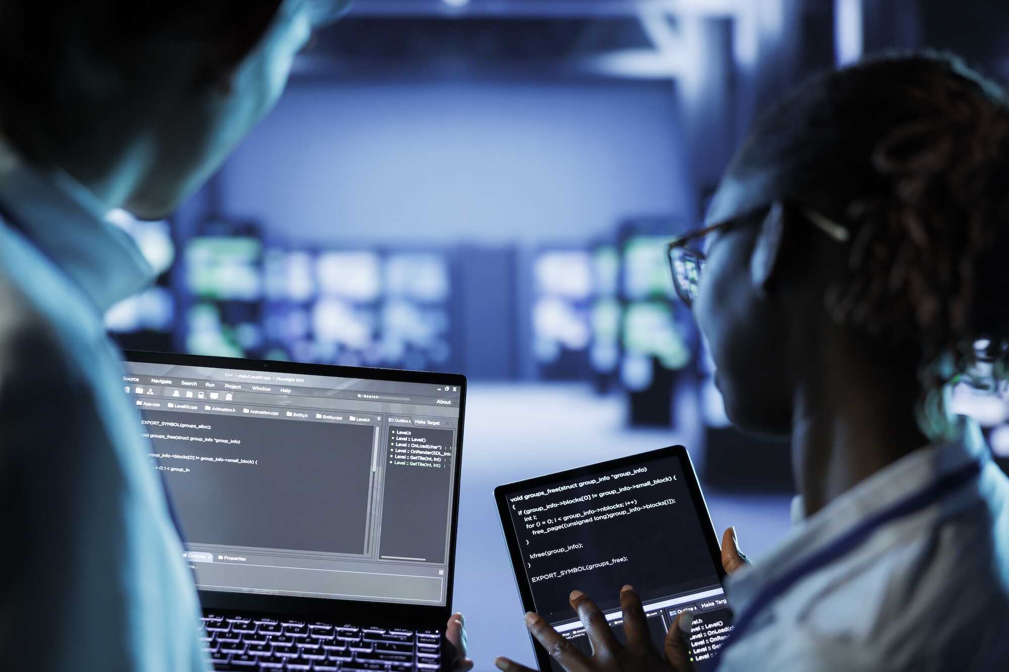 Two programmers in a dimly lit room working together, one holding a laptop and the other a tablet, both displaying computer code on their screens.