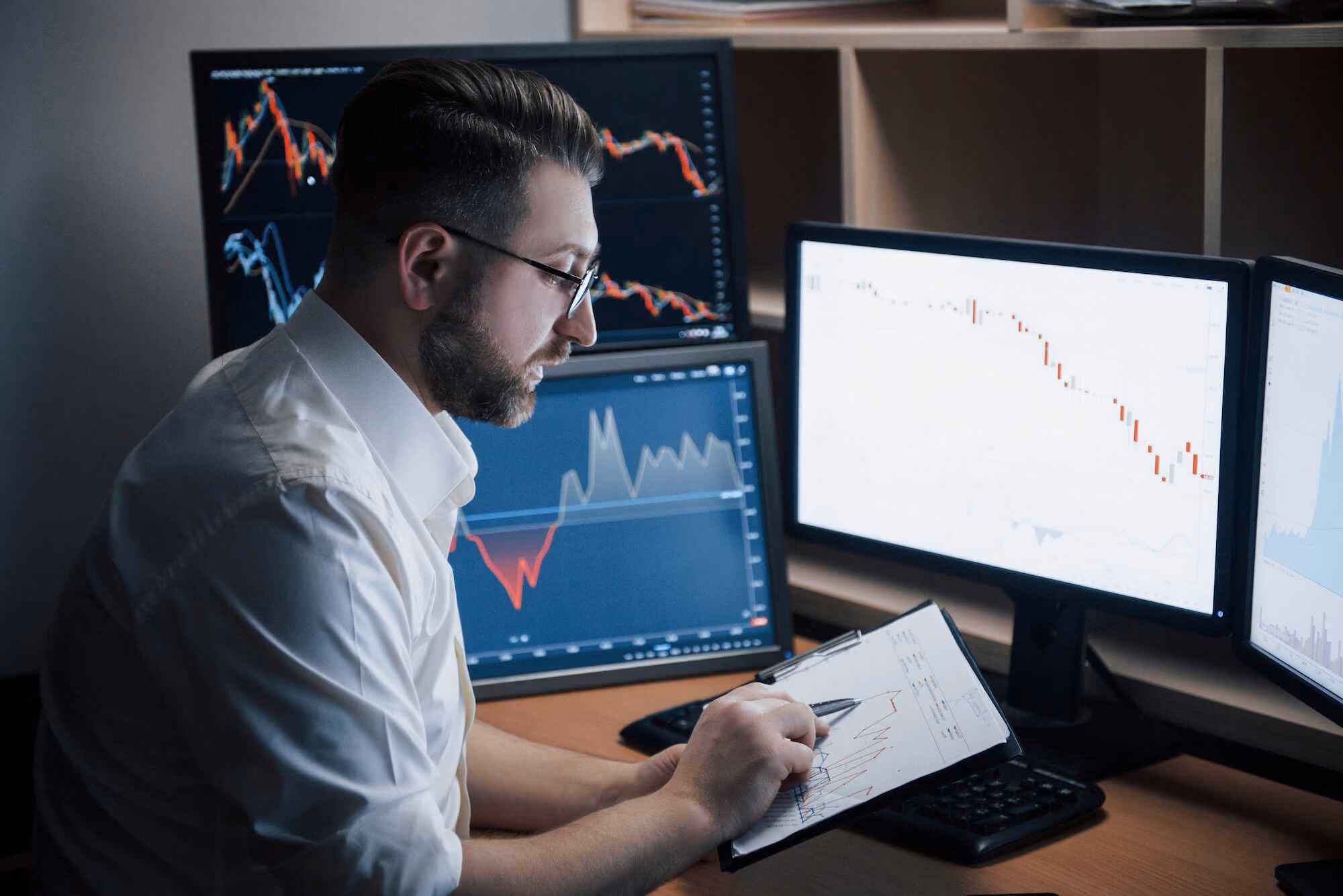 Man wearing glasses and a white shirt analyzing financial charts on multiple computer screens and a clipboard in a dimly lit office.