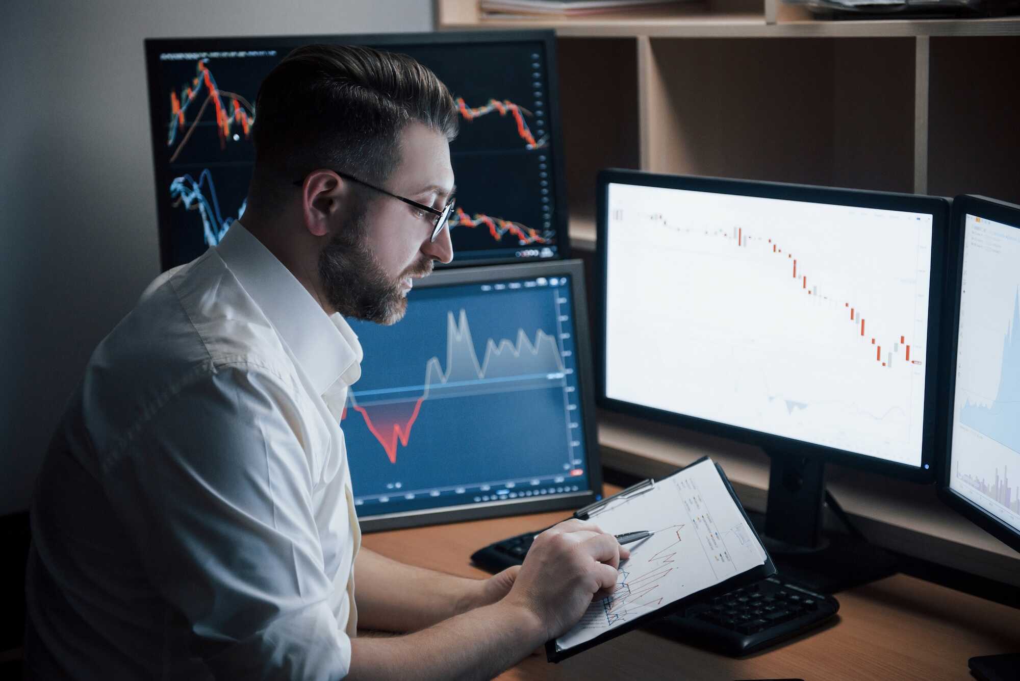 Man wearing glasses and a white shirt analyzing financial charts on multiple computer screens and a clipboard in a dimly lit office.