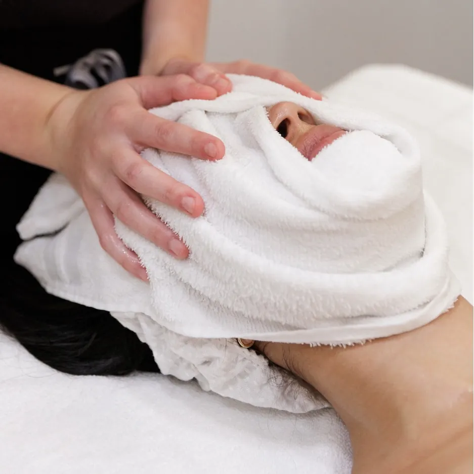 Esthetician wrapping a client's face in a warm towel during a facial