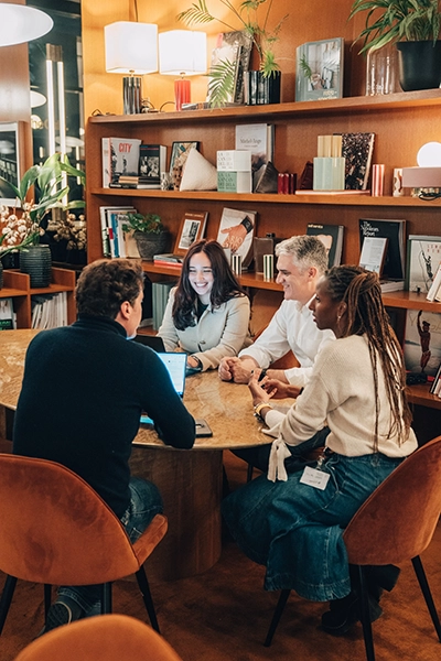 Un groupe diversifié de quatre personnes est assis autour d'une table en bois dans une pièce confortable et remplie de livres, engagés dans une discussion animée et positive.