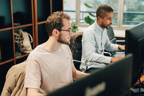 Deux hommes travaillent à des bureaux d'ordinateur dans un bureau. L'un porte des lunettes et une chemise rose, l'autre une chemise bleu clair. Ils semblent concentrés et engagés.