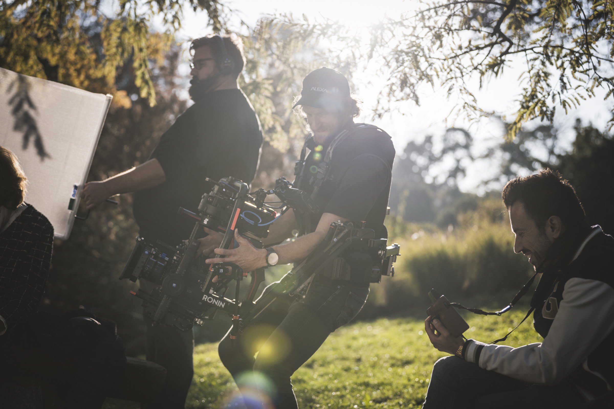 La troupe al lavoro in esterni: un operatore con rig stabilizzato Ronin, un pannello riflettente e il focus puller, in controluce
