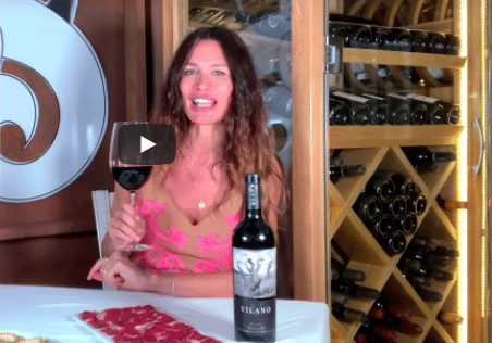 Fotograma de vídeo en el que Maria Zabay sostiene una copa de vino tinto junto a una botella de Vilano, delante de un botellero de madera