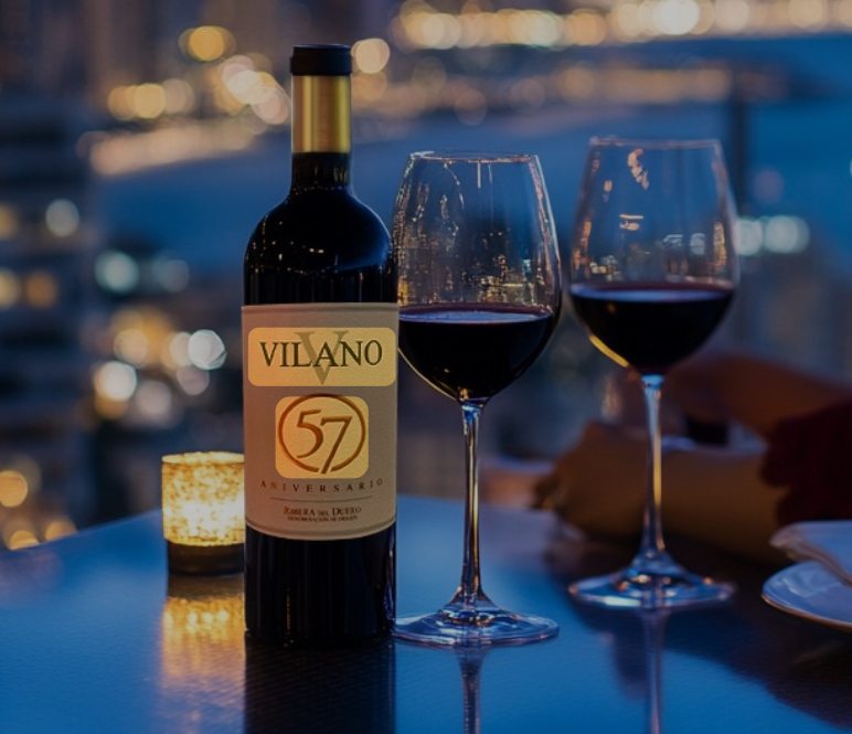 Botella de Vilano 57 Aniversario y dos copas de vino tinto sobre una mesa, con las luces de la ciudad de noche al fondo