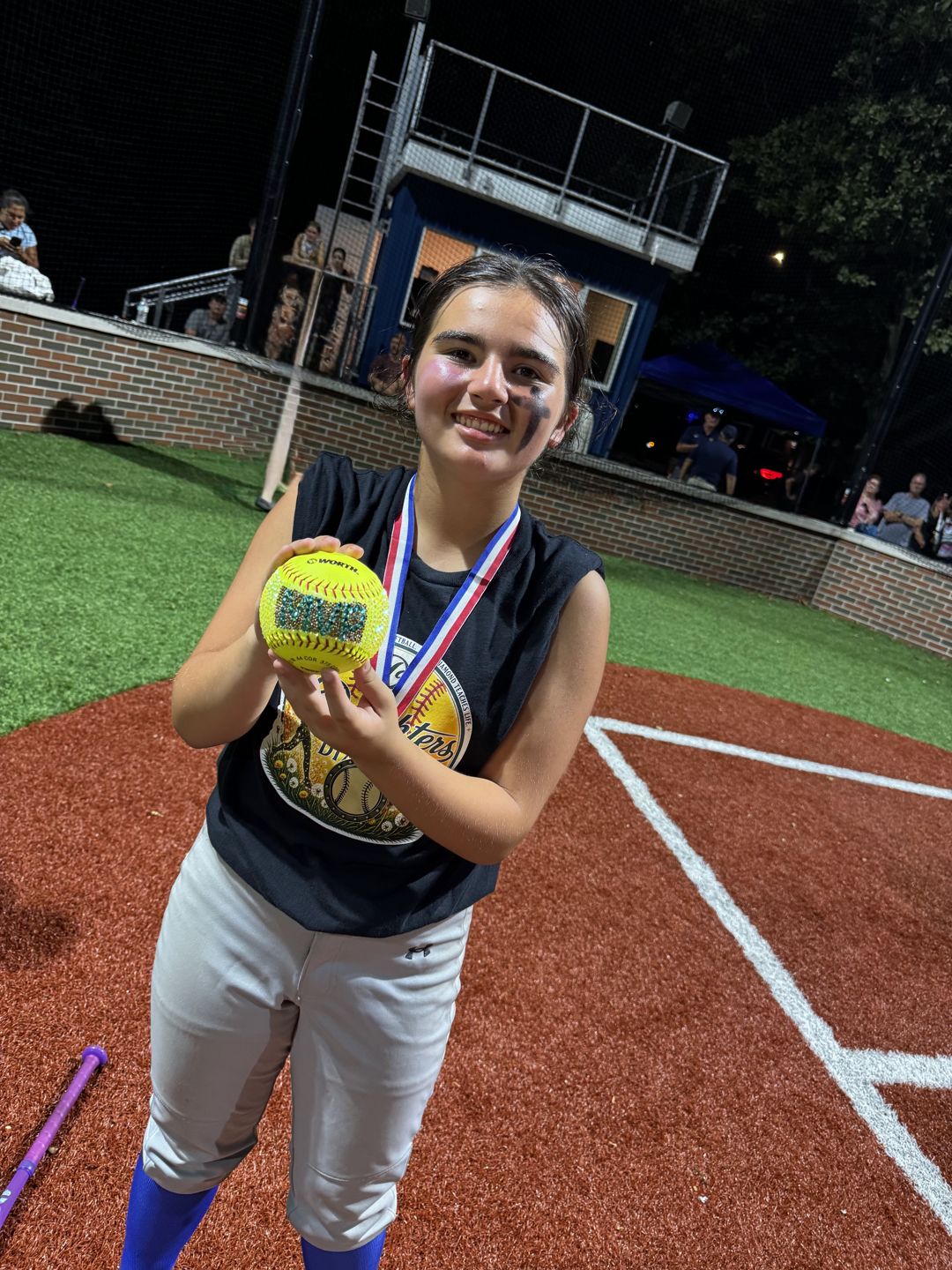 A player holds up a gold softball and her medal after the All-Star game under the lights.