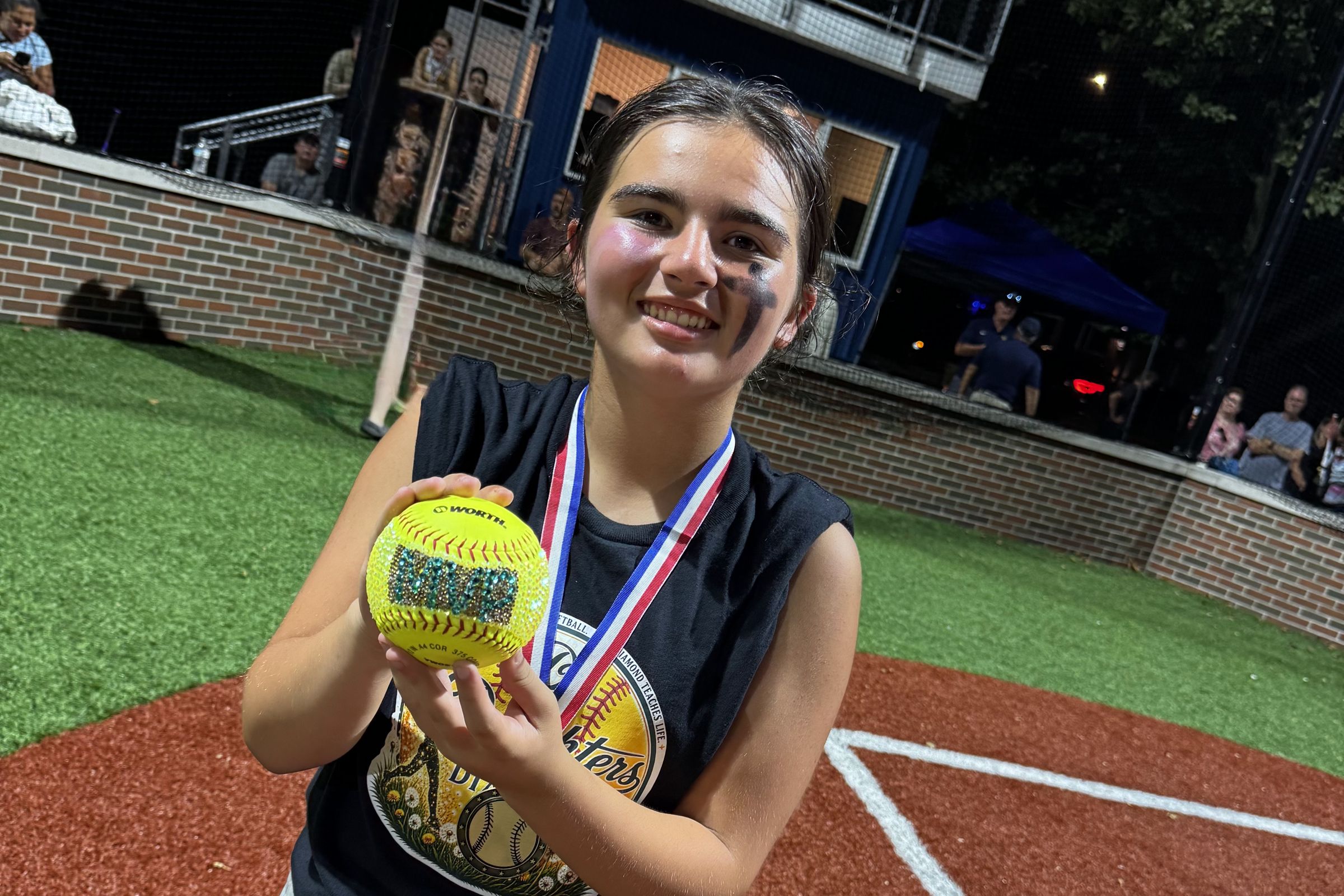 A player holds up a gold softball and her medal after the All-Star game under the lights.