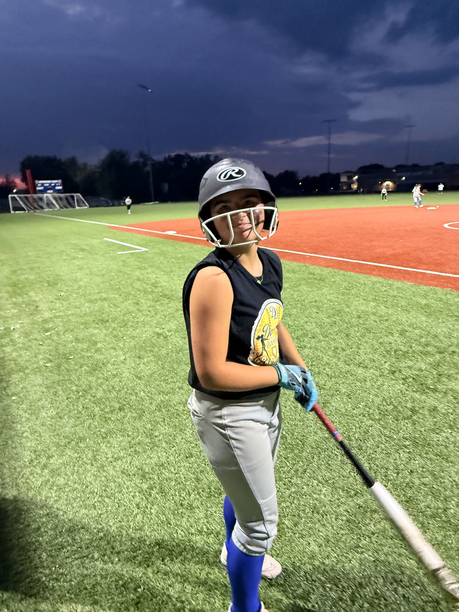 A player in a helmet and Daughters on the Diamond jersey smiles with her bat at dusk.