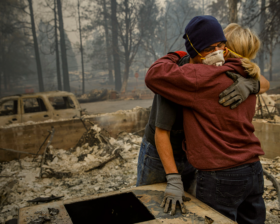 Two people in protective masks and gloves embrace amidst the charred remains of a forest.