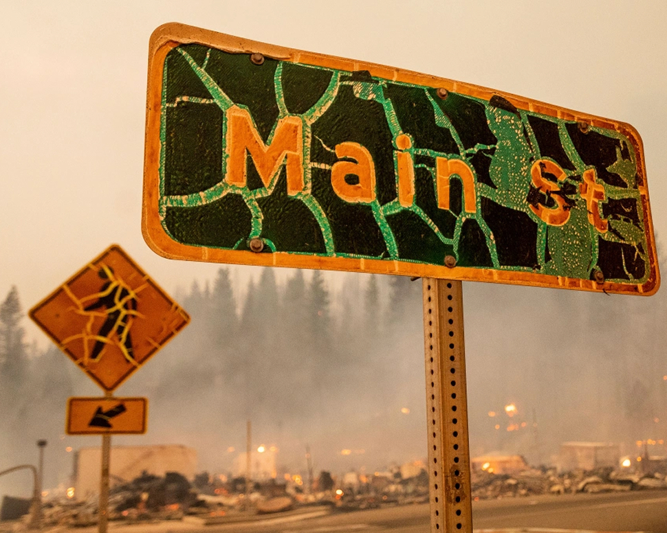 A damaged Main St sign stands in front of a hazy, smoke-filled background.