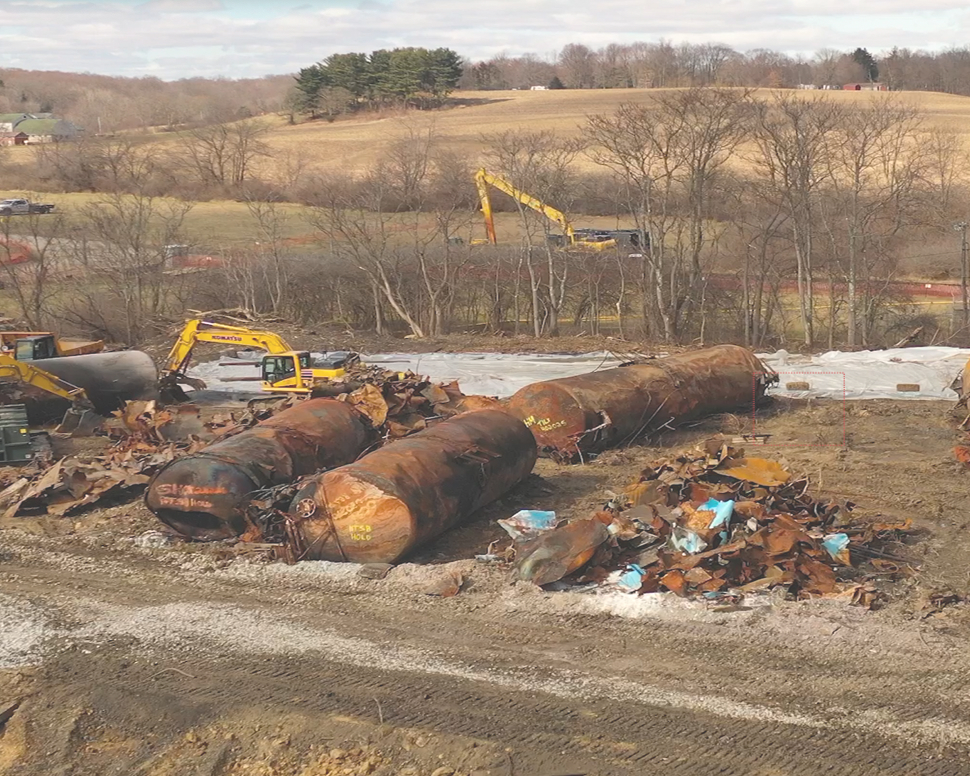 Construction site with rusted, dismantled fuel tanks, excavators, and debris on a dirt landscape.