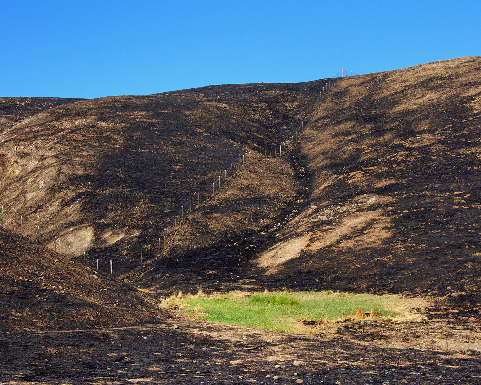 A landscape of charred hills with a narrow green patch in the center under a clear blue sky.