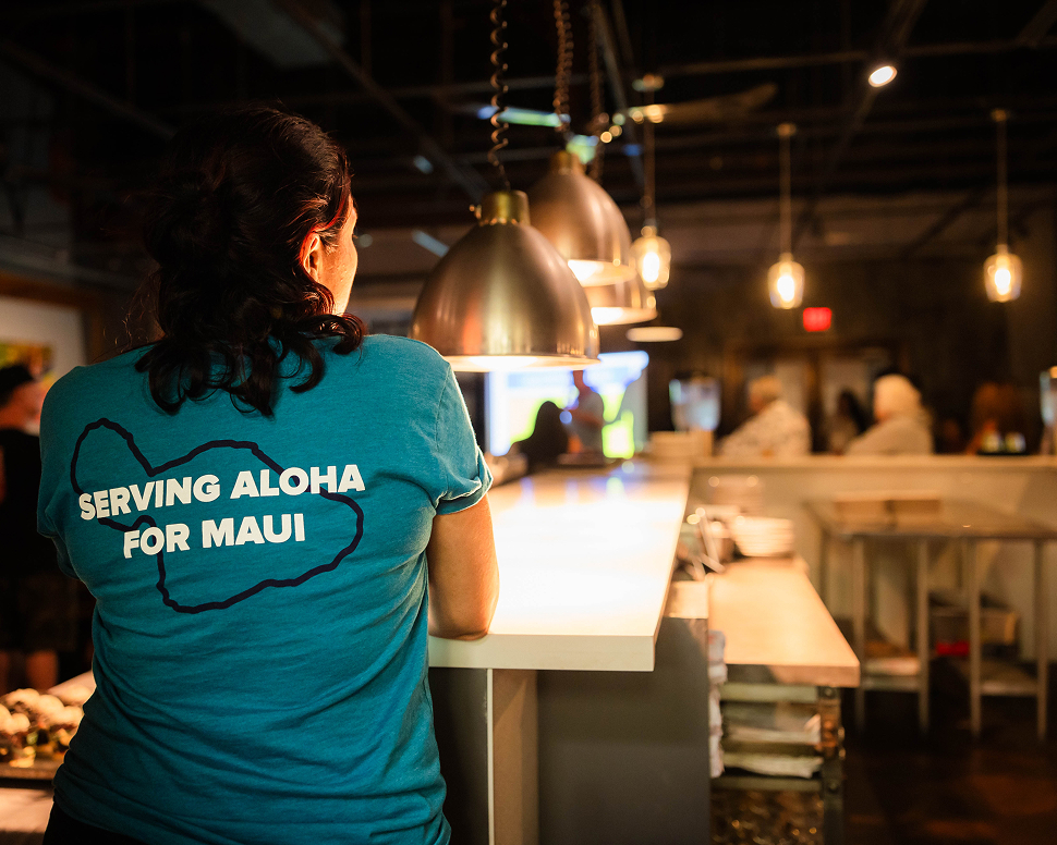 Person in a blue shirt reading Serving Aloha for Maui stands at a counter in a warmly lit restaurant.