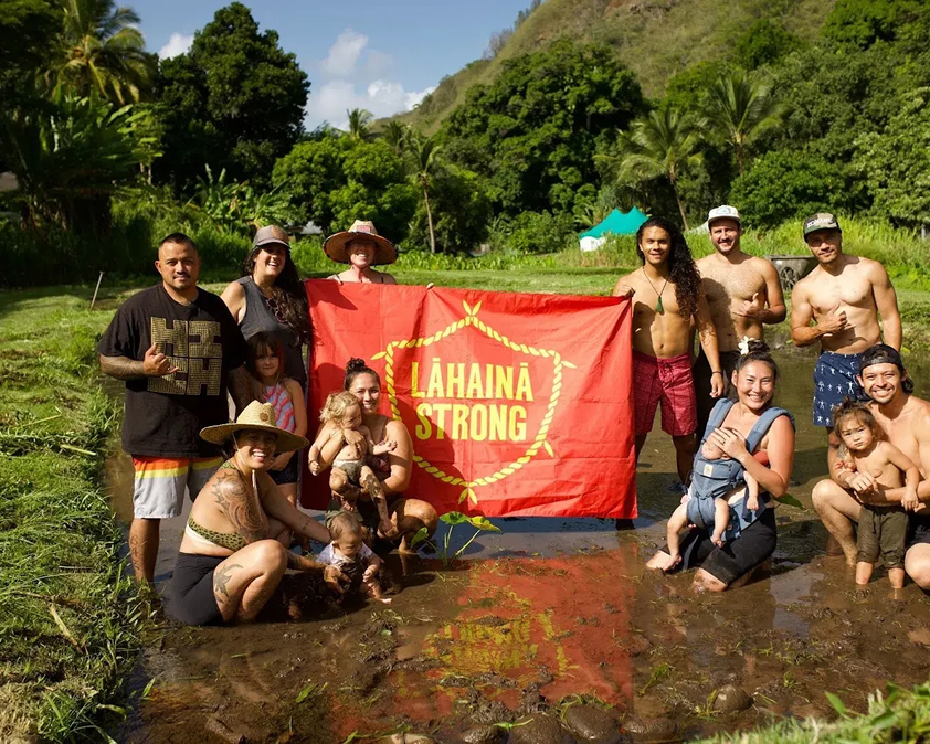 Group of people in a lush, tropical setting holding a red Lahaina Strong banner.