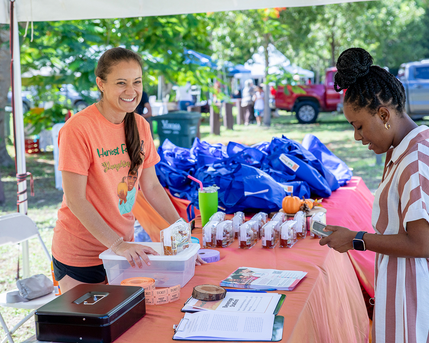 A woman at a booth with an orange tablecloth and blue bags smiles at another person.