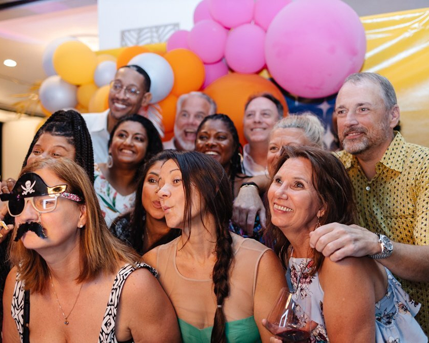 A group of people smiling and posing together at a party, with colorful balloons in the background.