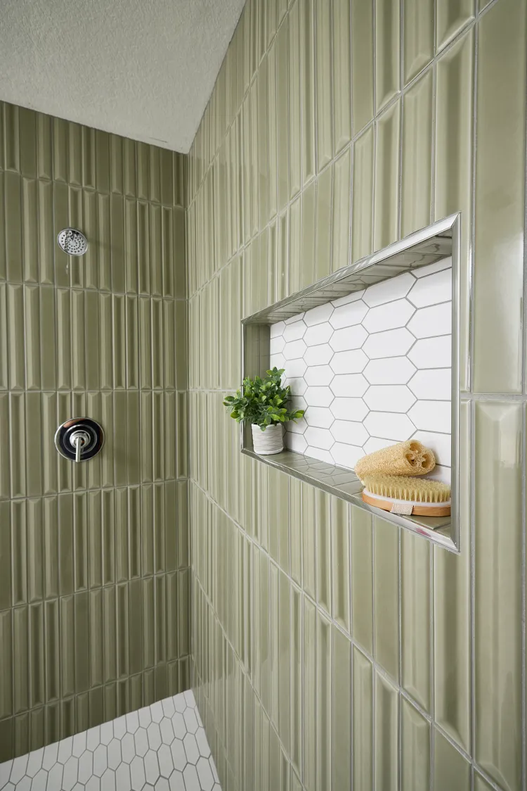 Shower with vertical olive-green tiles, white hexagonal floor tiles, and a niche with plant and bath brush.