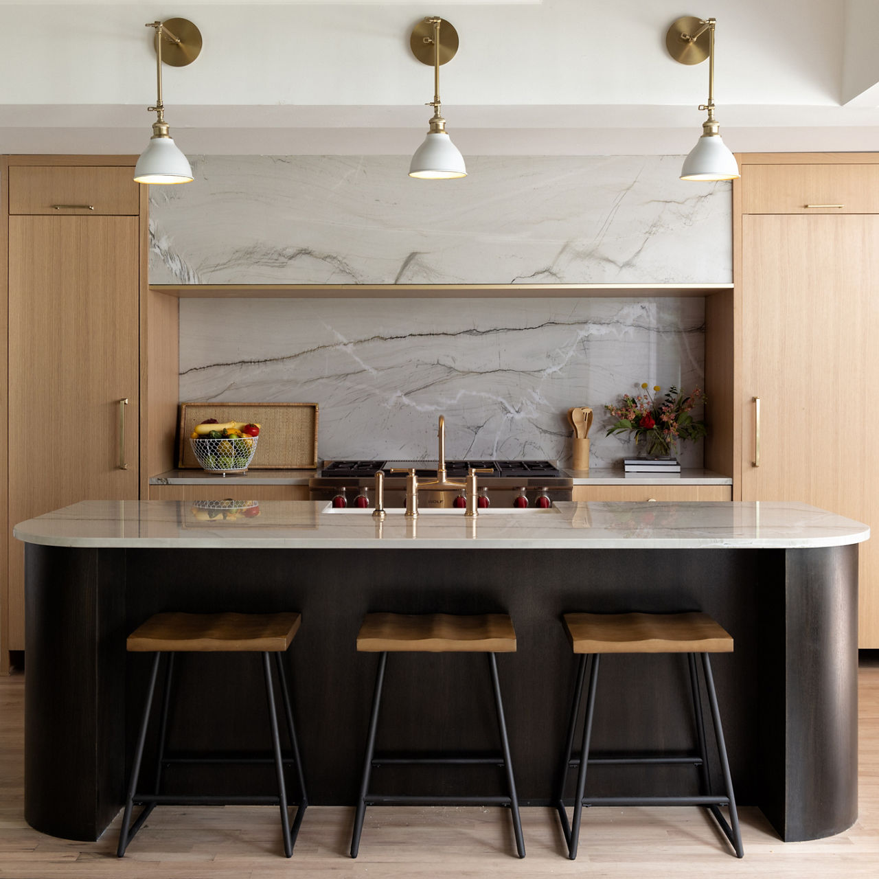 Modern kitchen with marble island, three stools, brass fixtures, wooden cabinets, and pendant lights.