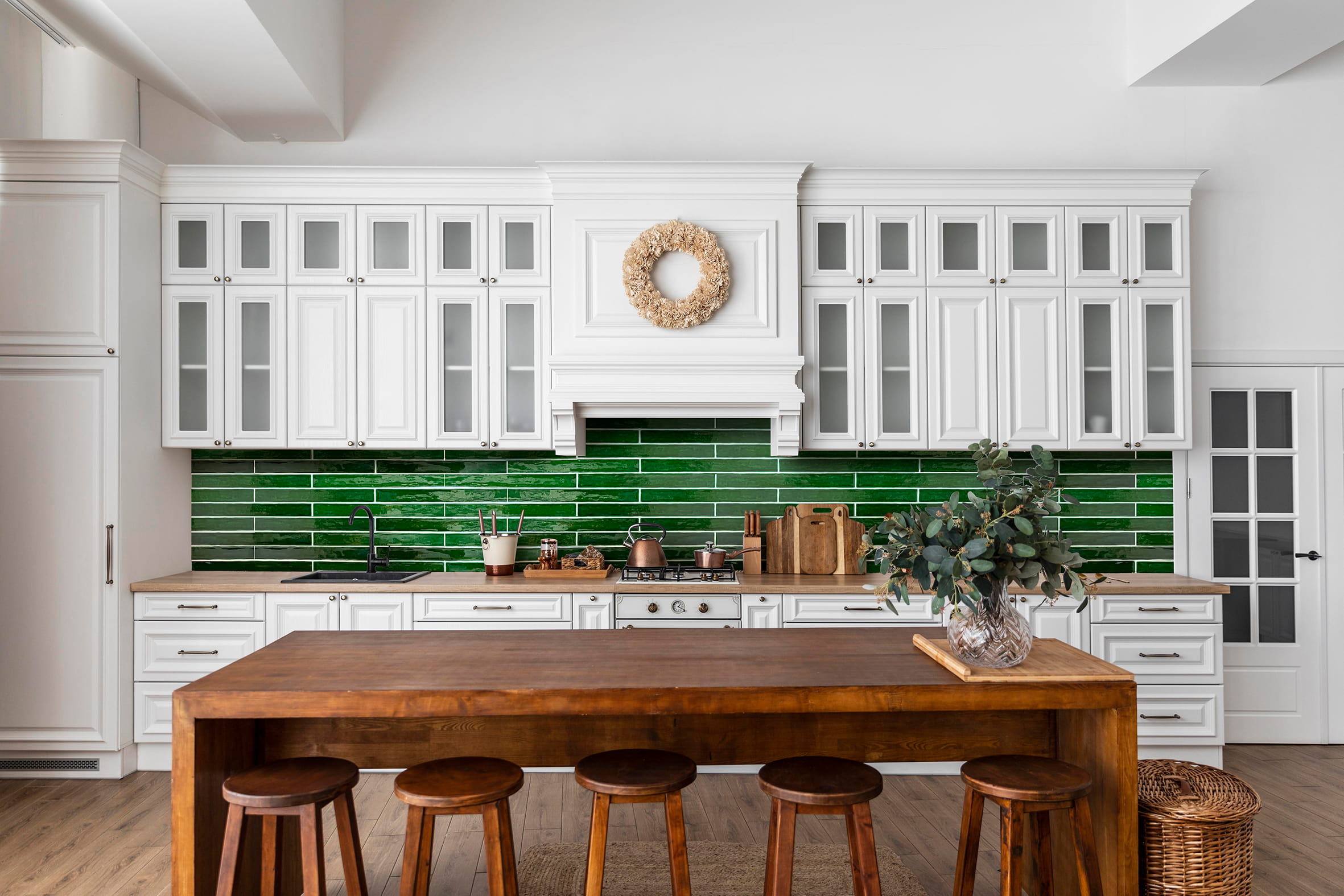 Modern kitchen with white cabinets, green tile backsplash, wooden island, stools, and vase with greenery.