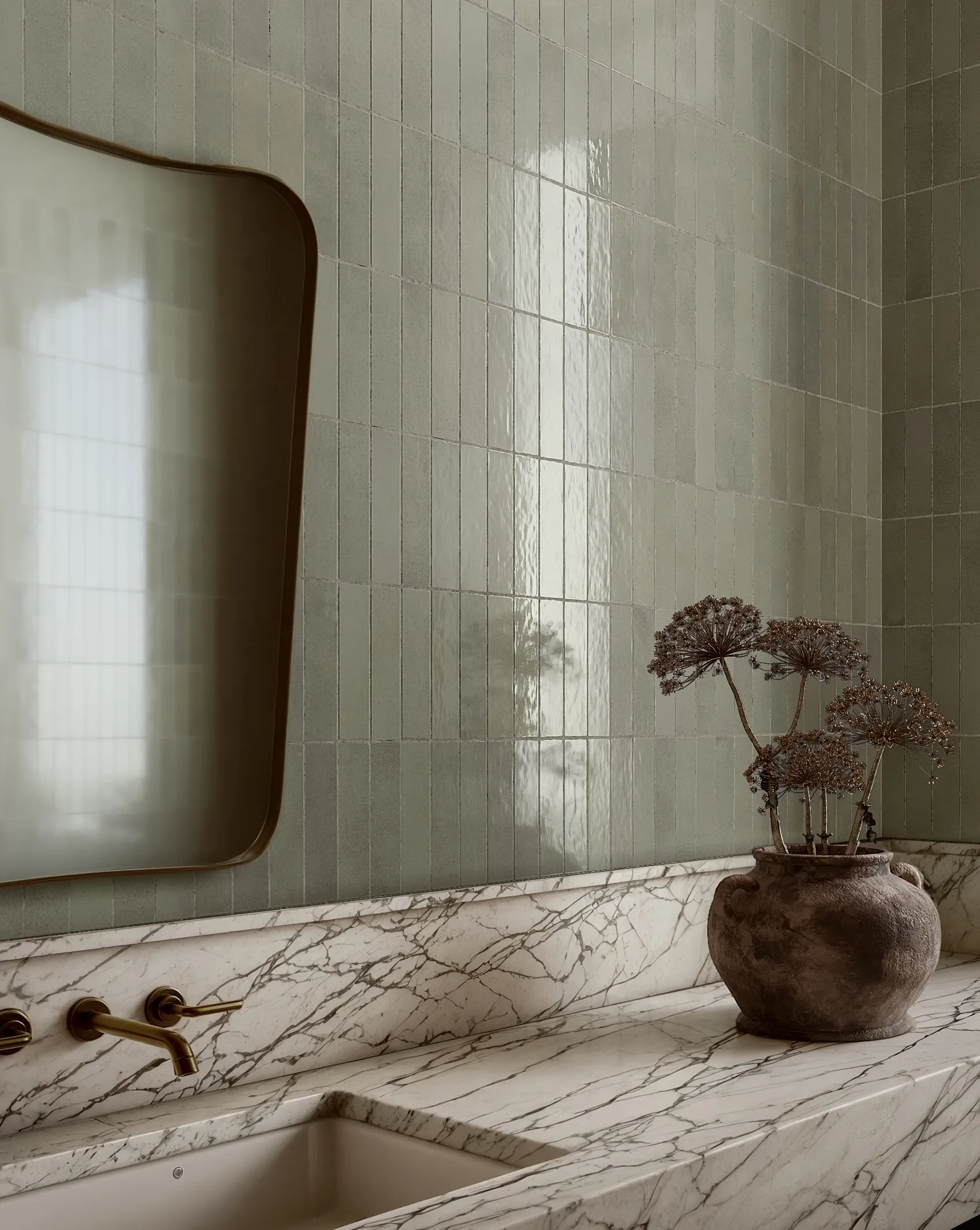 Bathroom sink with marble counter, brass faucet, muted green tiled walls, and a rustic vase with dried flowers.