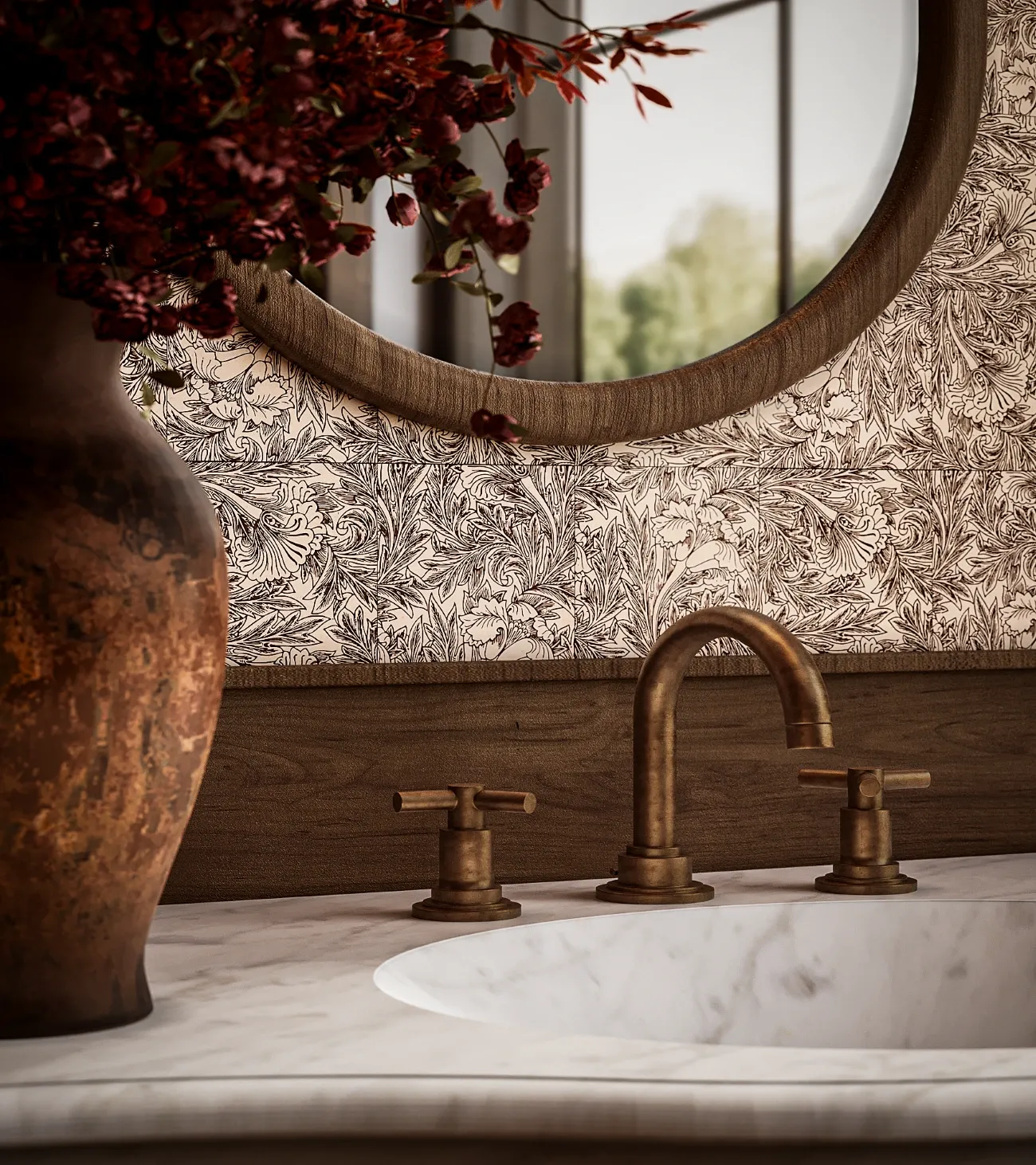 Marble sink with bronze faucet, floral patterned wallpaper, wooden framed round mirror, and vase with red flowers.