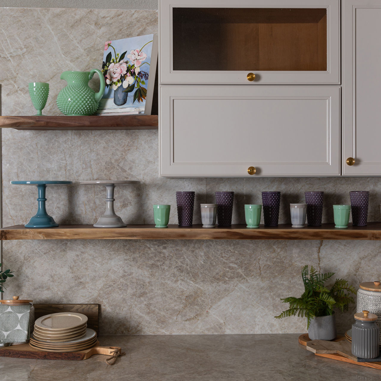 Kitchen shelves with green and purple glasses, cake stands, floral painting, plates, plants, and jars.