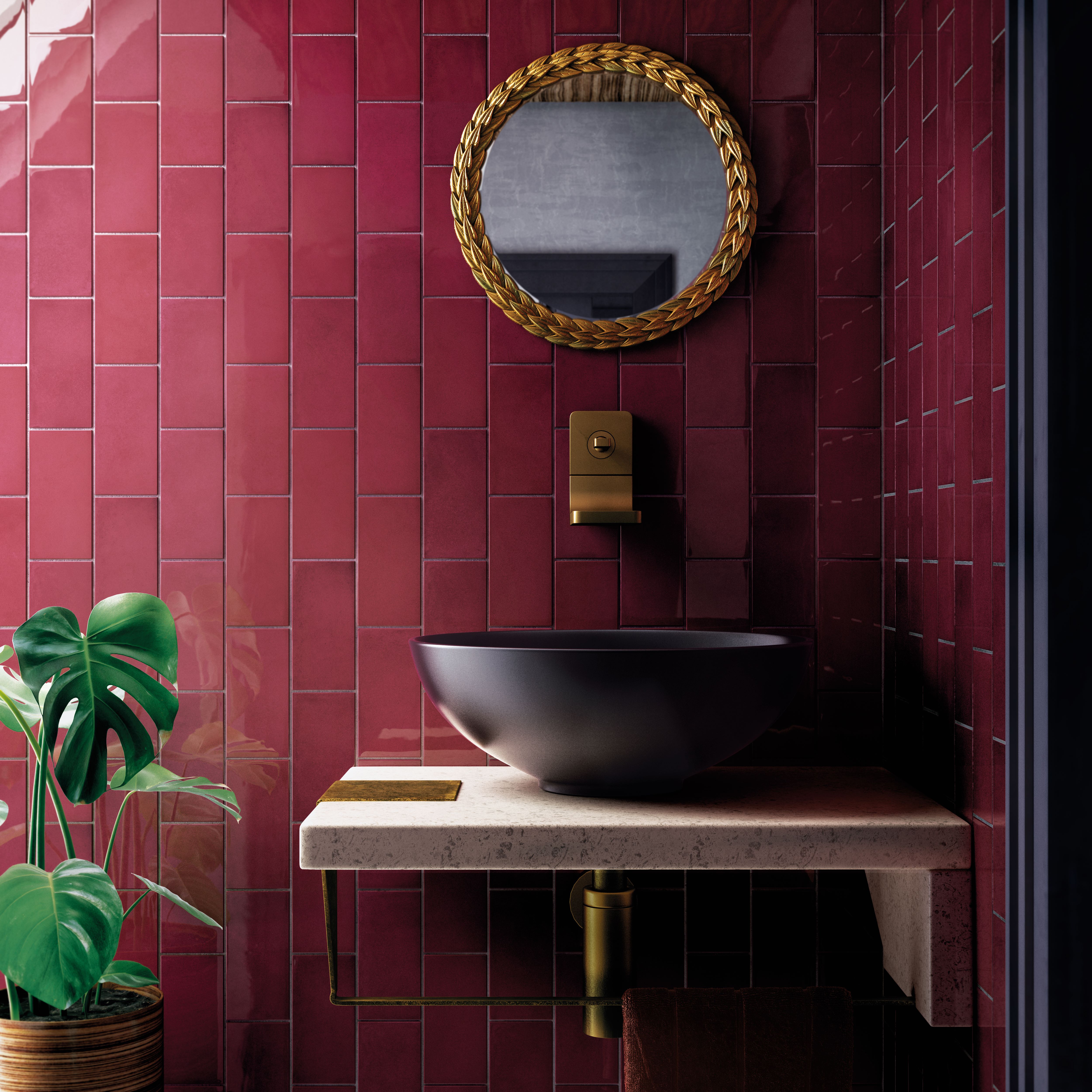 Modern bathroom sink with black bowl, gold faucet, round mirror, and burgundy tile wall, plus green plant.