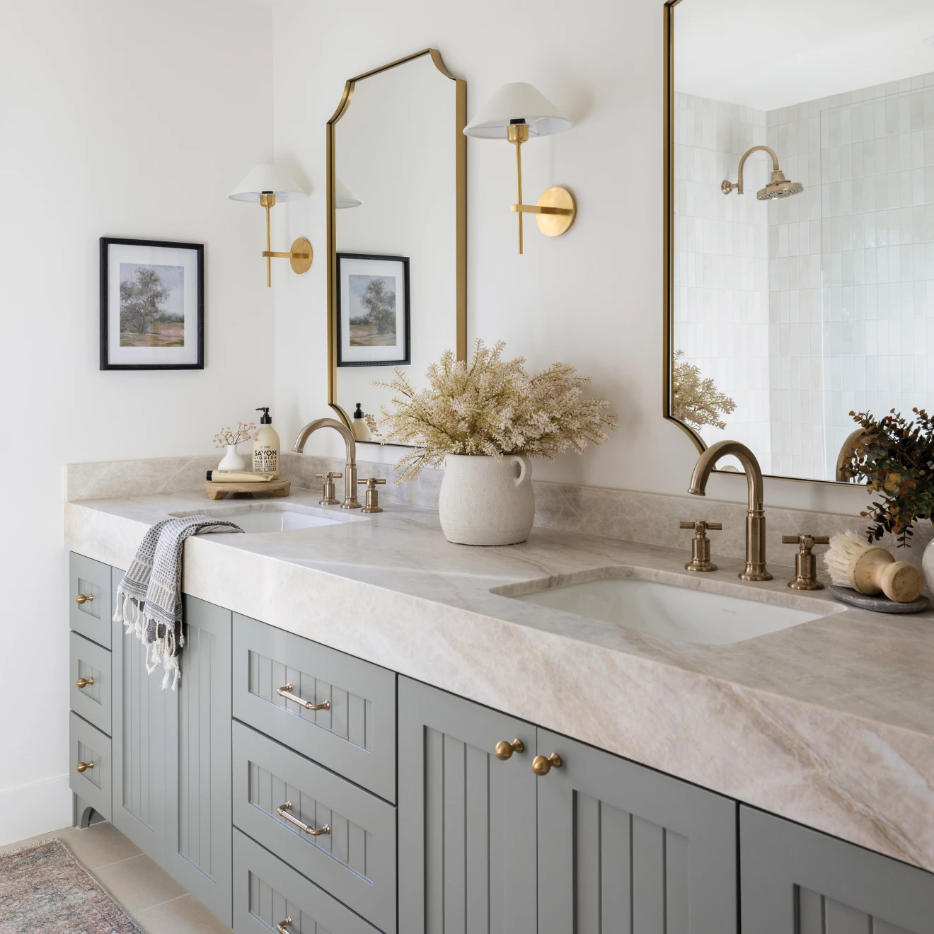 Modern double bathroom vanity with gray cabinets, marble countertop, gold fixtures, mirrors, and decorative plants.