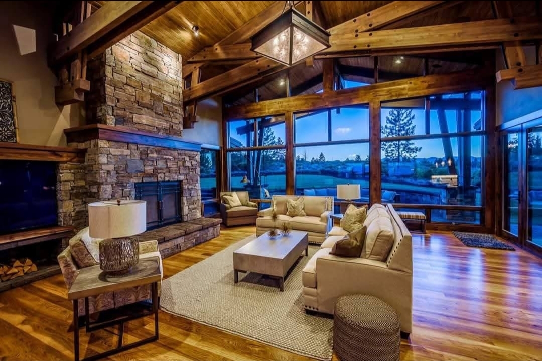 Spacious living room with large stone fireplace, wooden beams, sofas, and floor-to-ceiling windows showing outdoor view.