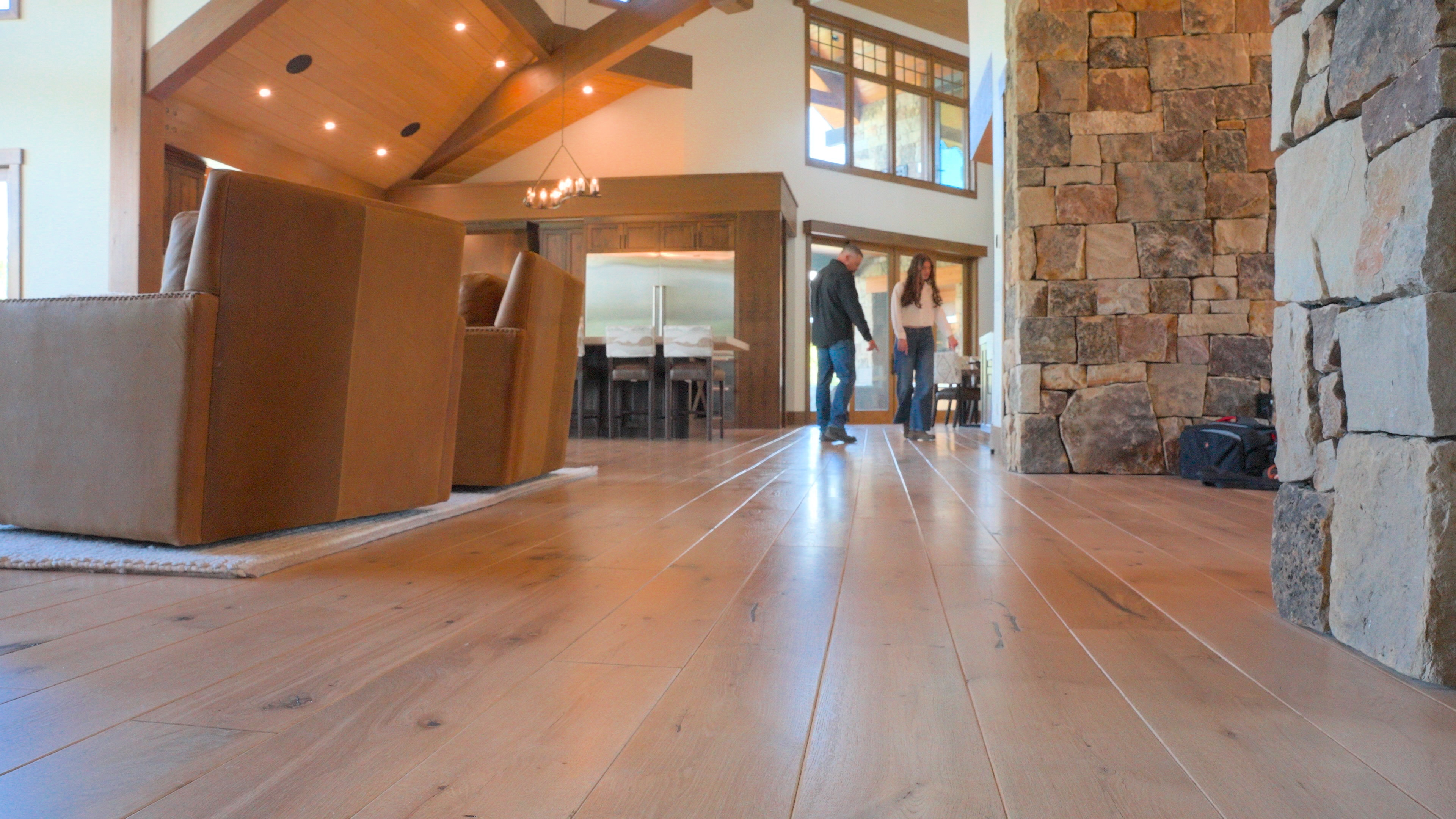 Two people standing in a modern living room with wooden floors, stone walls, and leather chairs.
