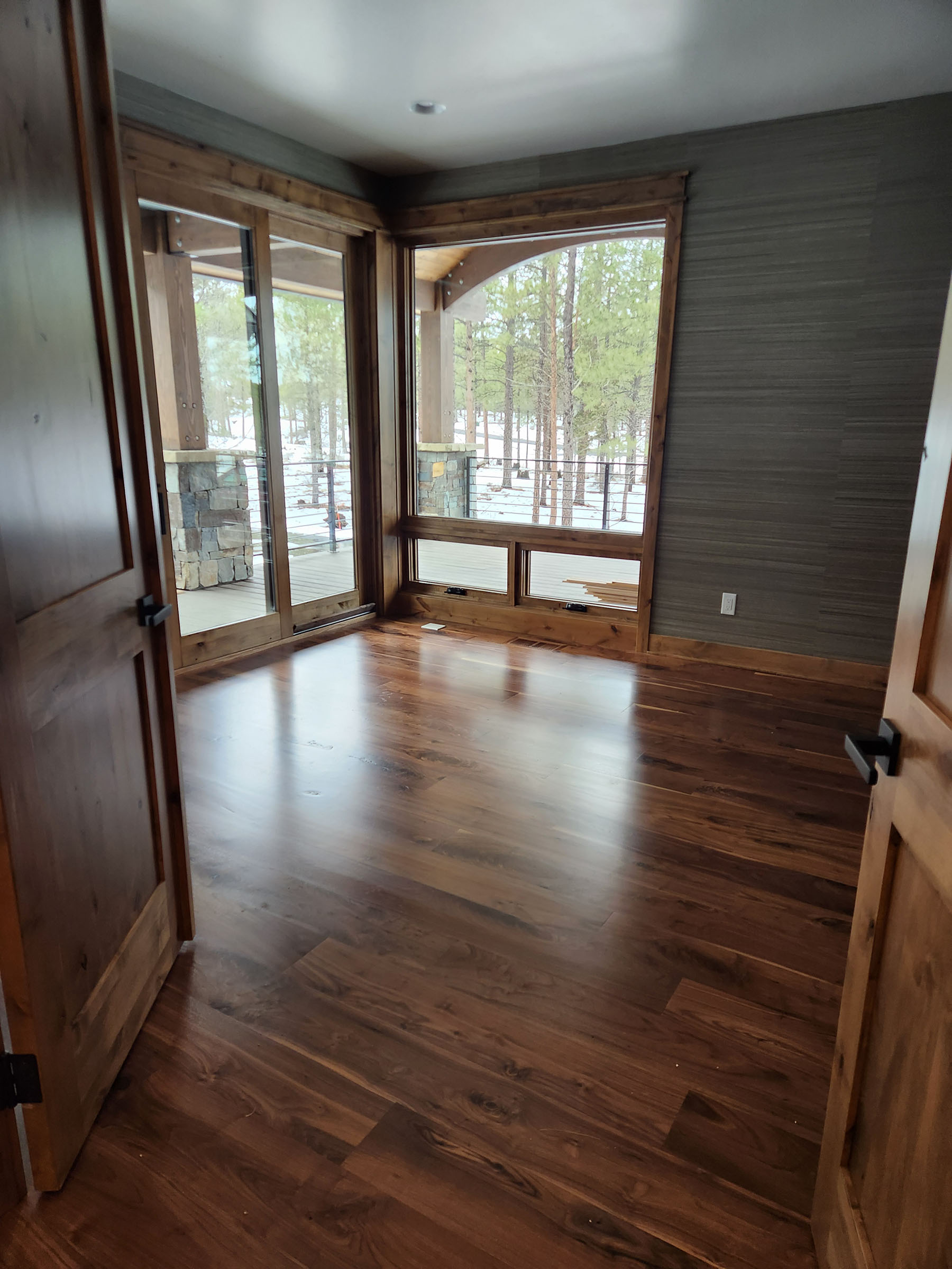 Empty room with wood floor, wooden doors, large windows, and an outdoor view of snow and trees.