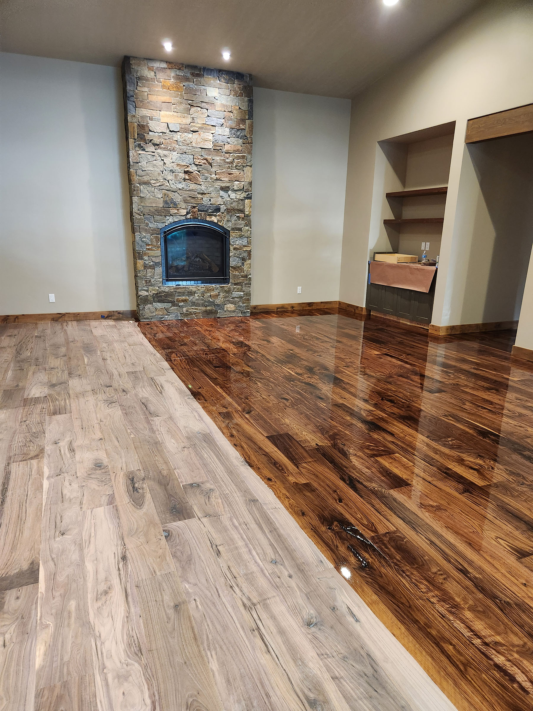 Room with unfinished and finished wood floors, stone fireplace, beige walls, and built-in shelves.