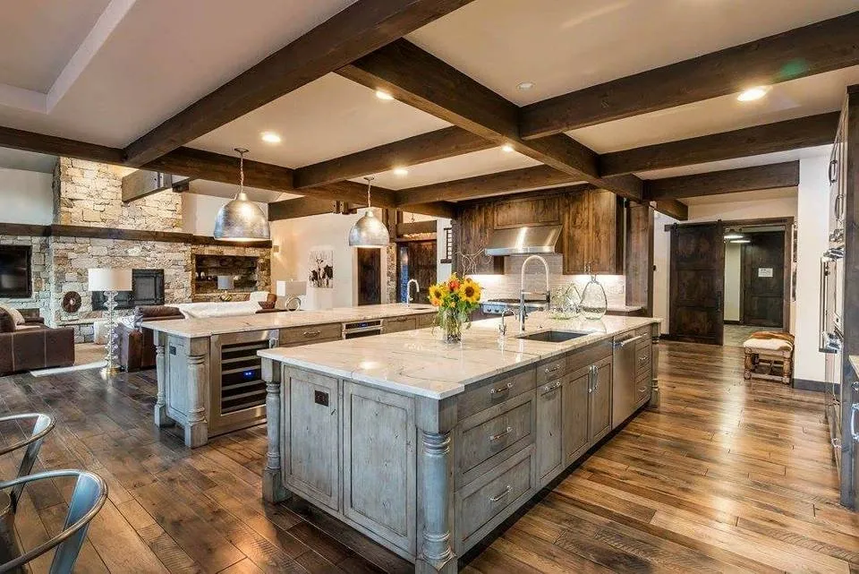 Spacious modern kitchen with two large islands, wooden beams, hardwood floors, and stone fireplace in background.