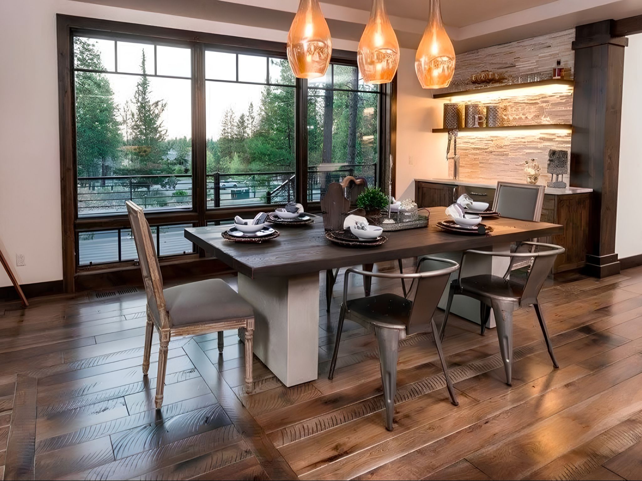 Modern dining room with wooden floor, dark dining table, various chairs, pendant lights, and large windows.