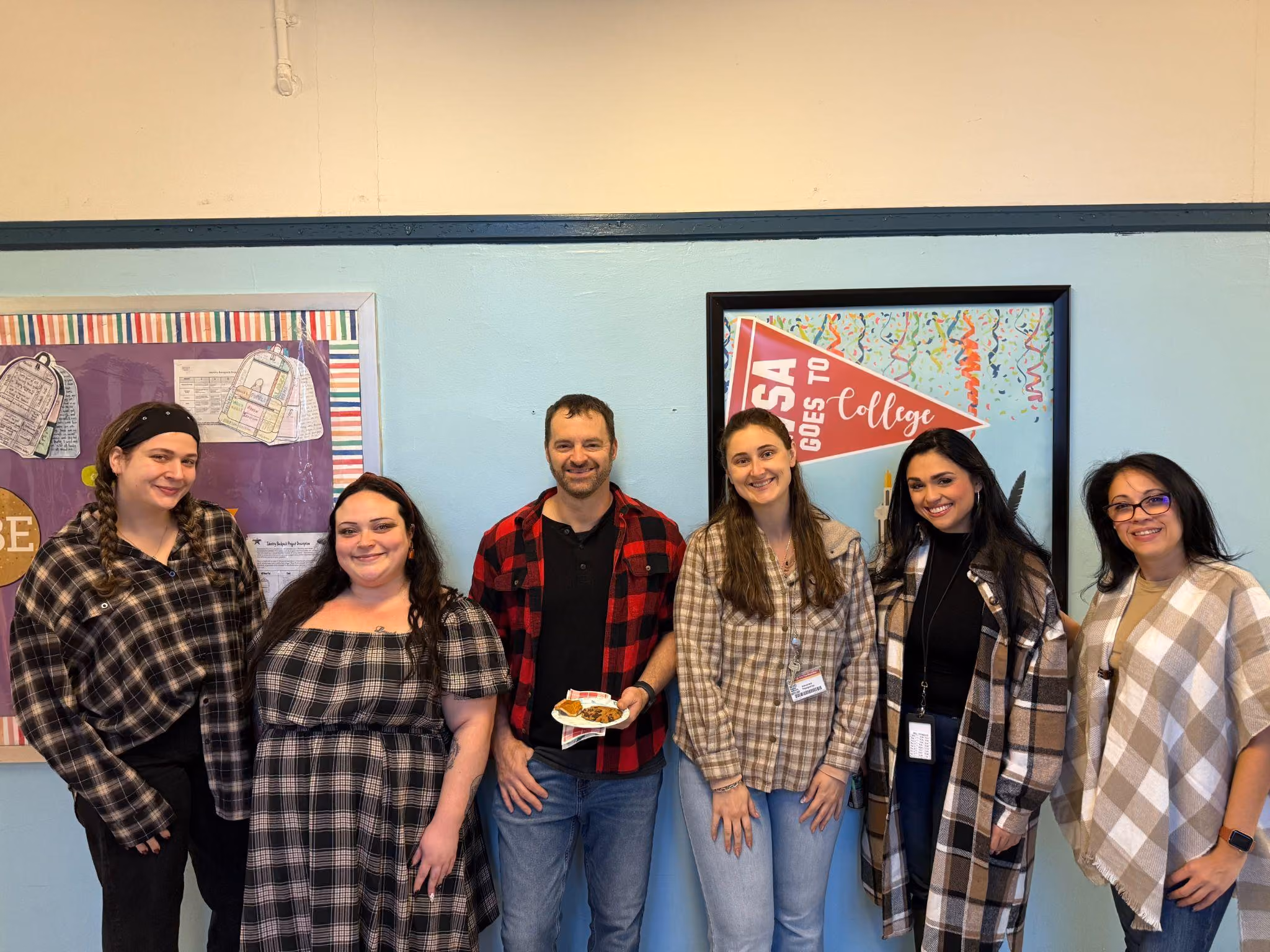 Six staff members posing together near a college bulletin board